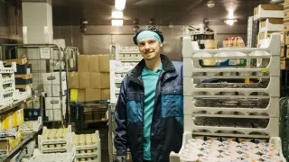 Henkilö seisoo kylmävarastossa jugurttipurkkien ympärillä ja hymyilee.