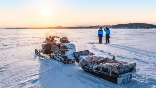 Valokuvassa moottorikelkka ja kaksi ihmistä auringonnousussa jäätyneellä järvellä. 